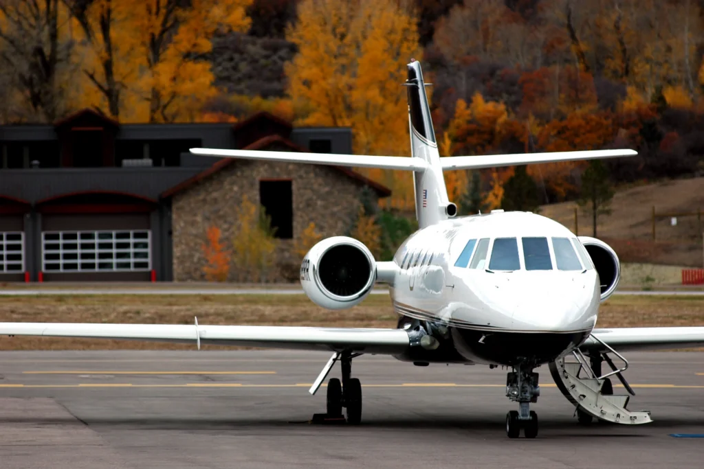 A white private jet with an open stairway is parked on an airport runway, with autumn trees and a building in the background.
