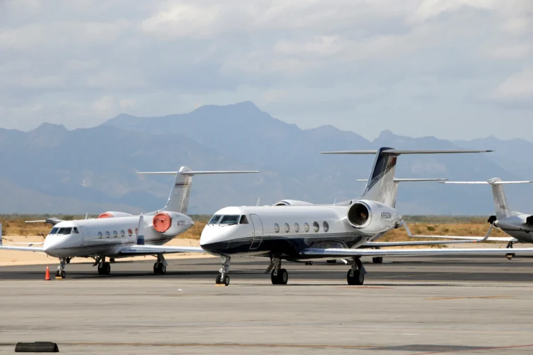Two private jets are parked on an airport tarmac with mountains visible in the background under a partly cloudy sky.