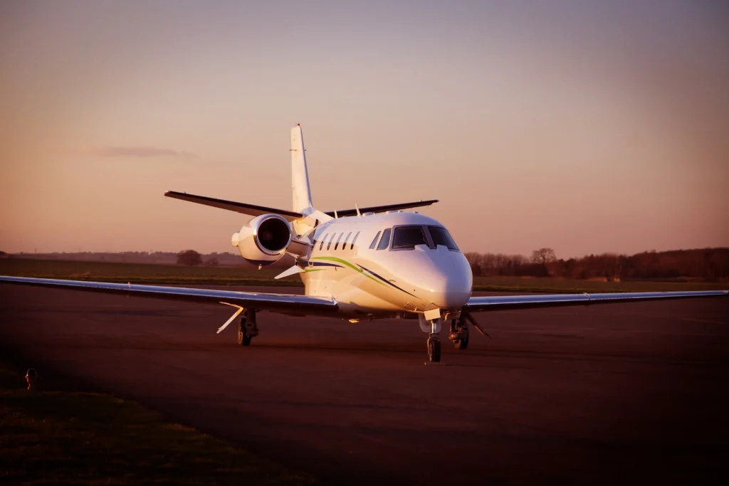 A white private jet is parked on an airport runway at sunset, with trees and open fields visible in the background.