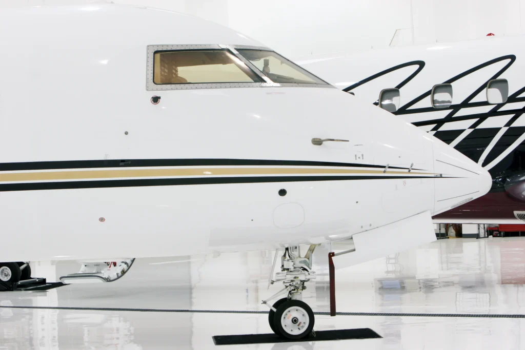 Close-up view of the nose and landing gear of a white private jet inside a hangar, with part of another jet visible in the background.