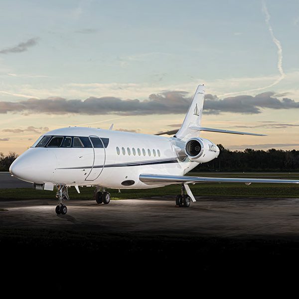 White private jet parked on an airport tarmac at sunset, with trees and a cloudy sky in the background.
